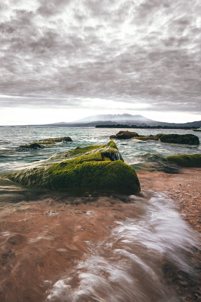Stunning view of moss-covered boulders and a cloudy sky over the ocean at sunset in Lombok, Indonesia.